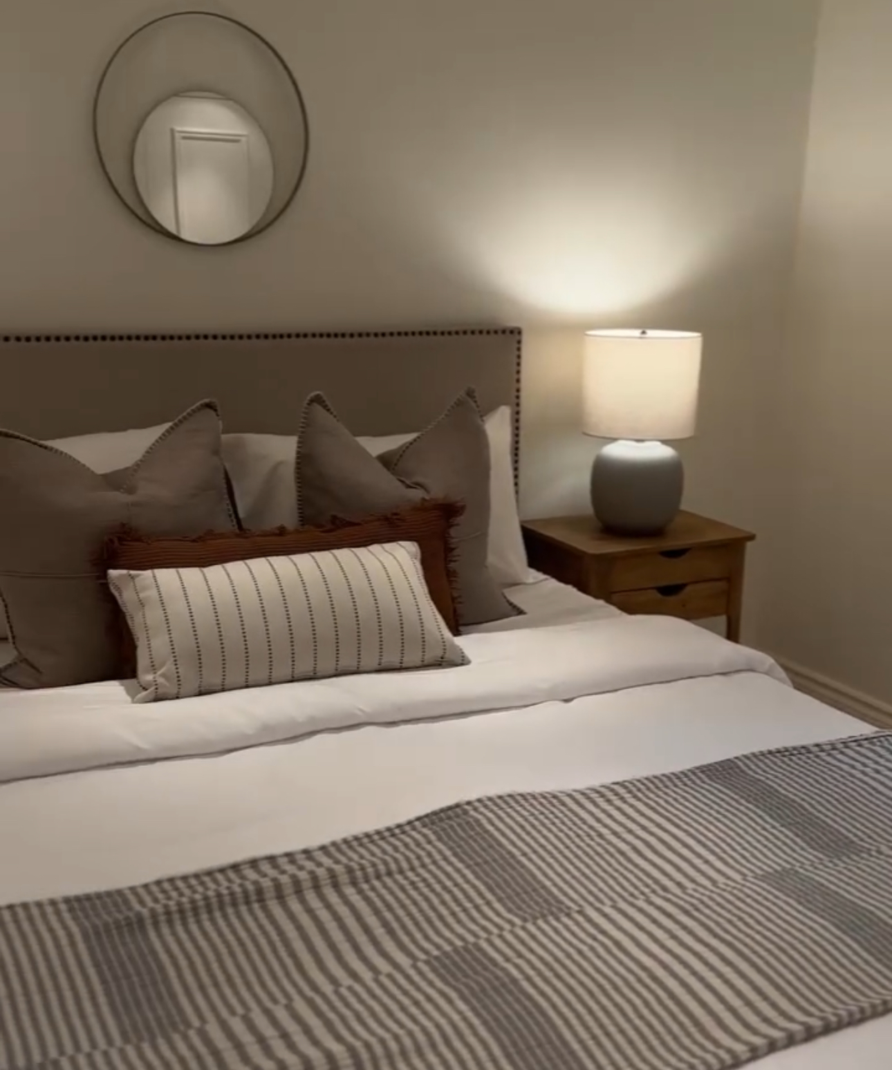 A staged bedroom with an upholstered headboard, white linens, decorative pillows and a lit bedside lamp