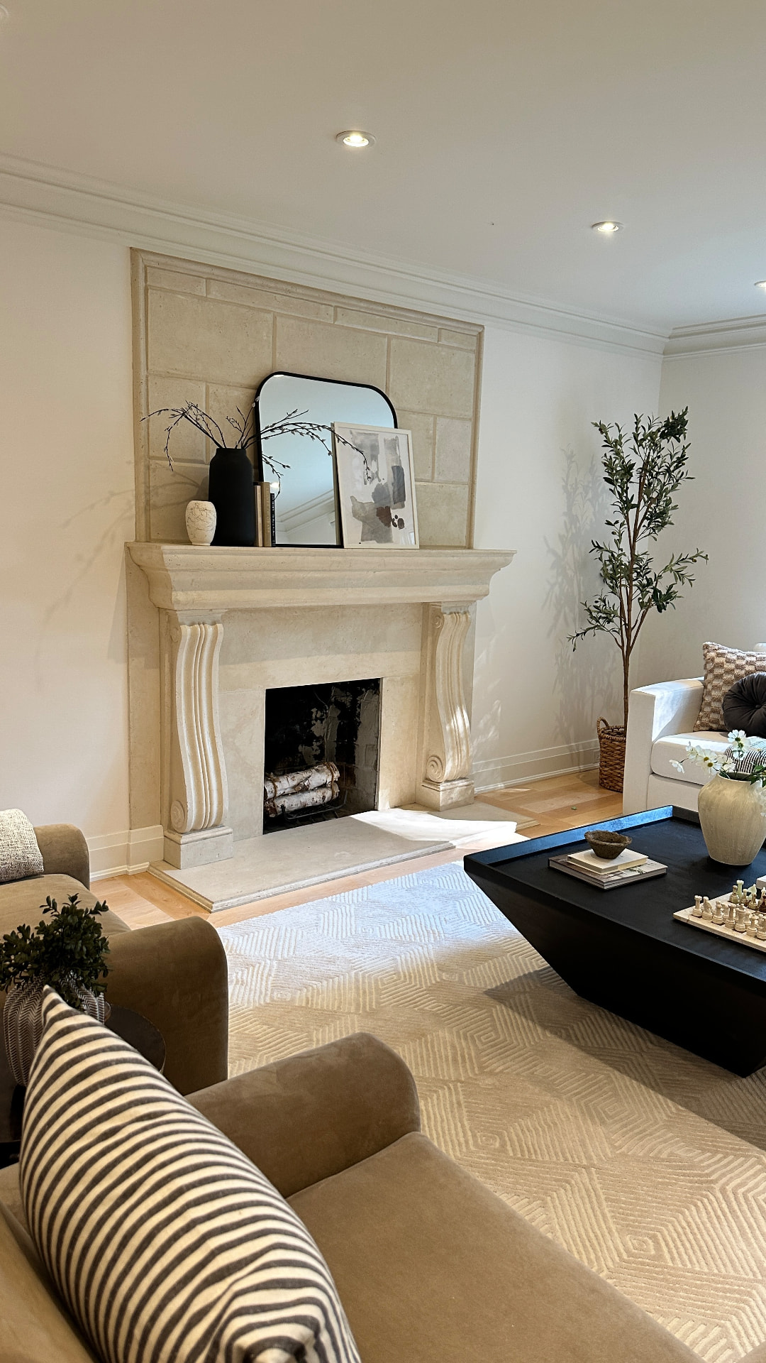 A staged living room with a carved stone fireplace, styled mantel, white sofa, potted olive tree and two tan armchairs around a low black coffee table