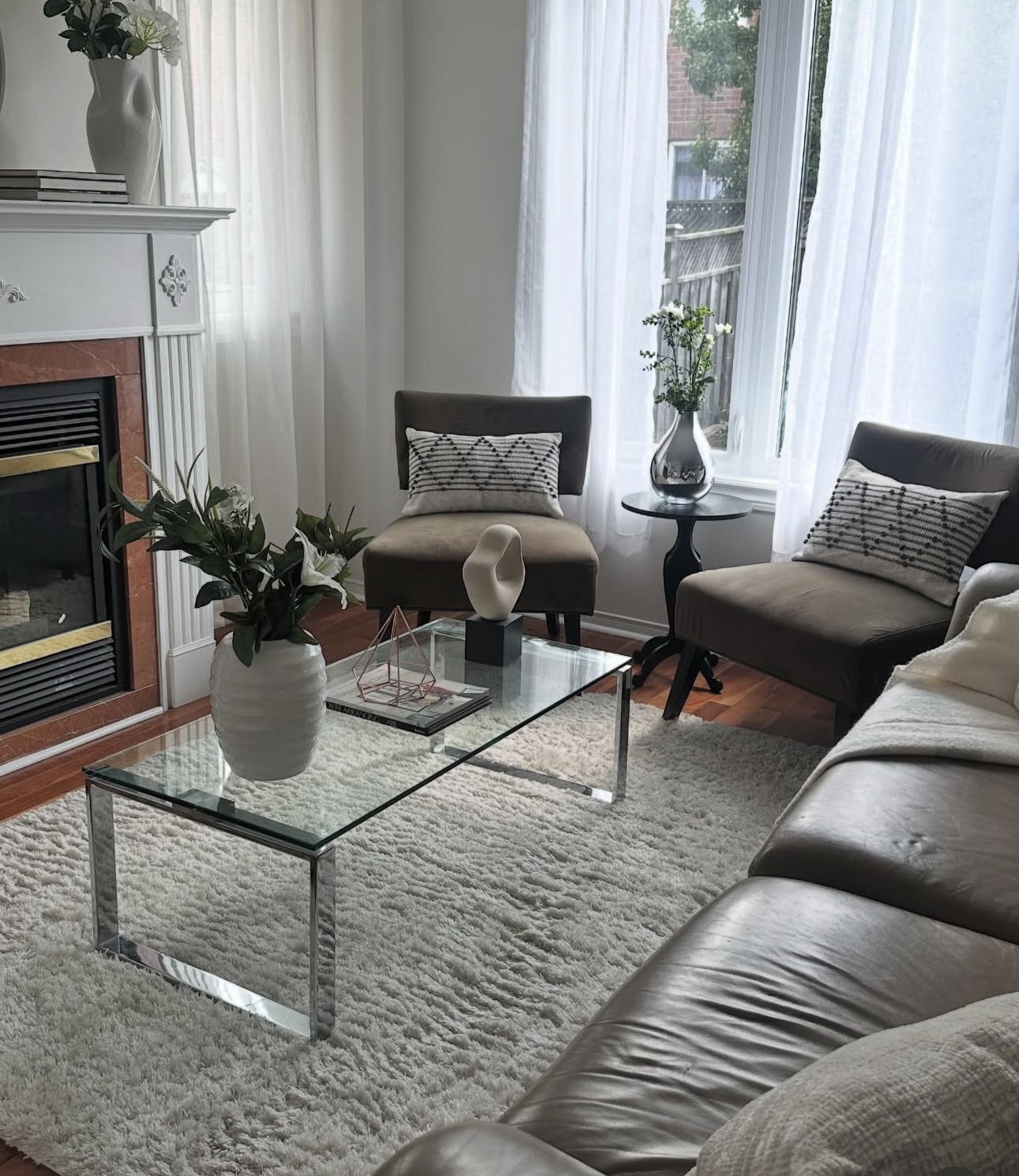 A staged living room with a white fireplace mantel, two accent armchairs, sheer curtains and a glass coffee table on a cream rug